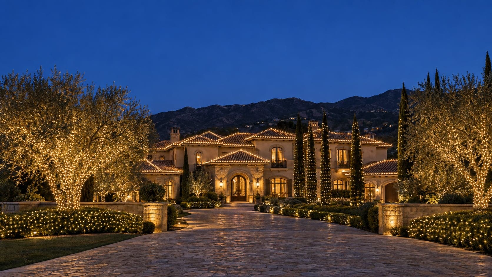 A Mediterranean estate at dusk with warm white lights on the roofline, olive trees and hedges